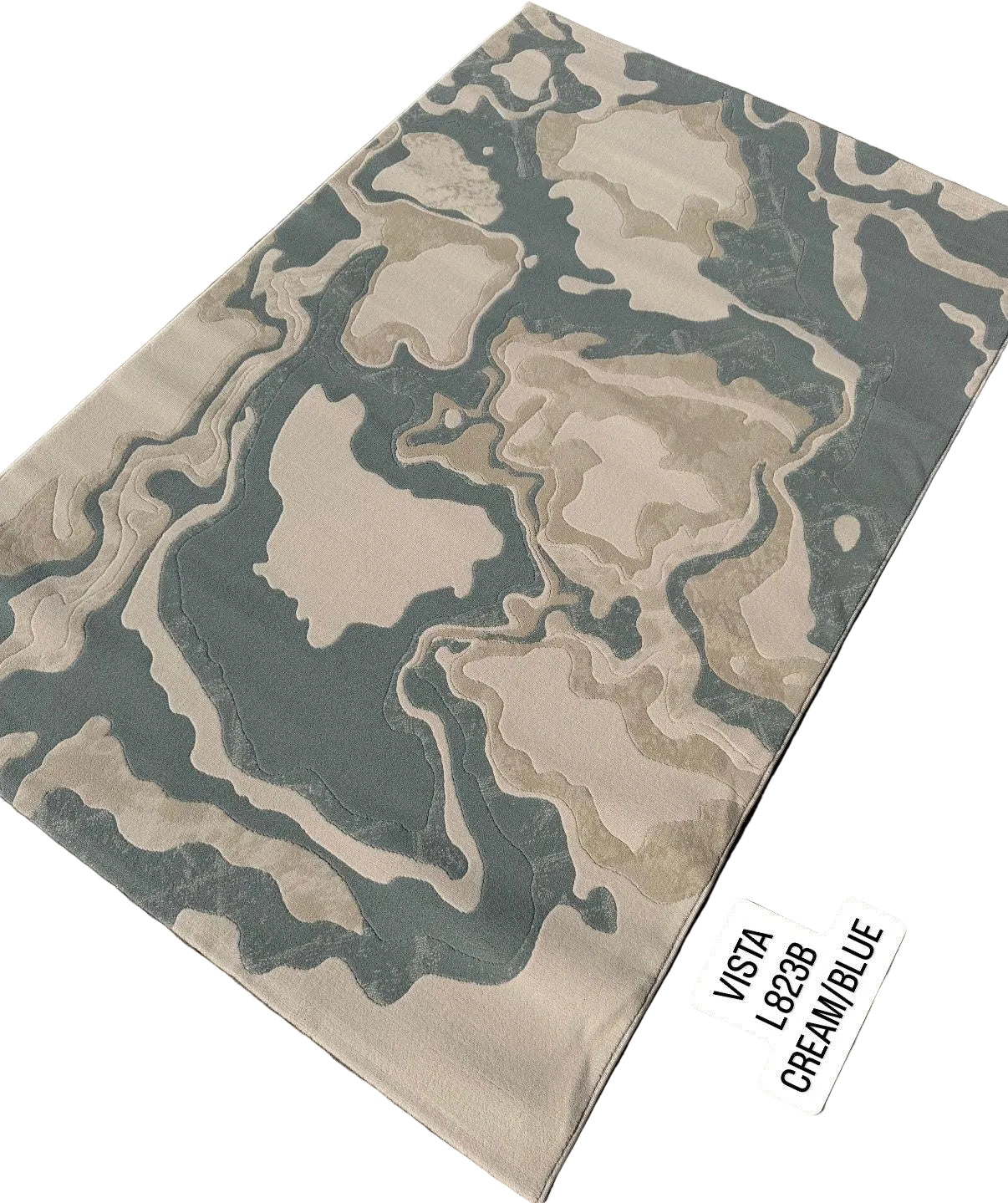 A rectangular area rug with an abstract, fluid topographic pattern in cream, beige, and muted blue-gray, shown laid out on a gray paved surface. Two white stickers label the colorway 'VISTA 1923 CREAM/BLUE'.