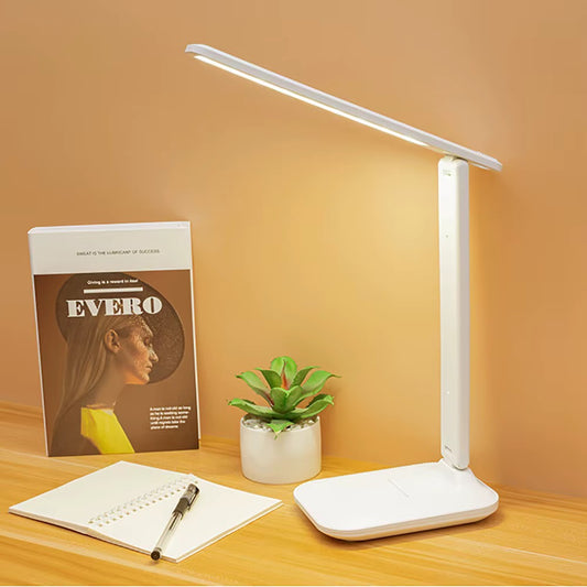 White desk lamp on a wooden surface with a book, plant, and notebook.